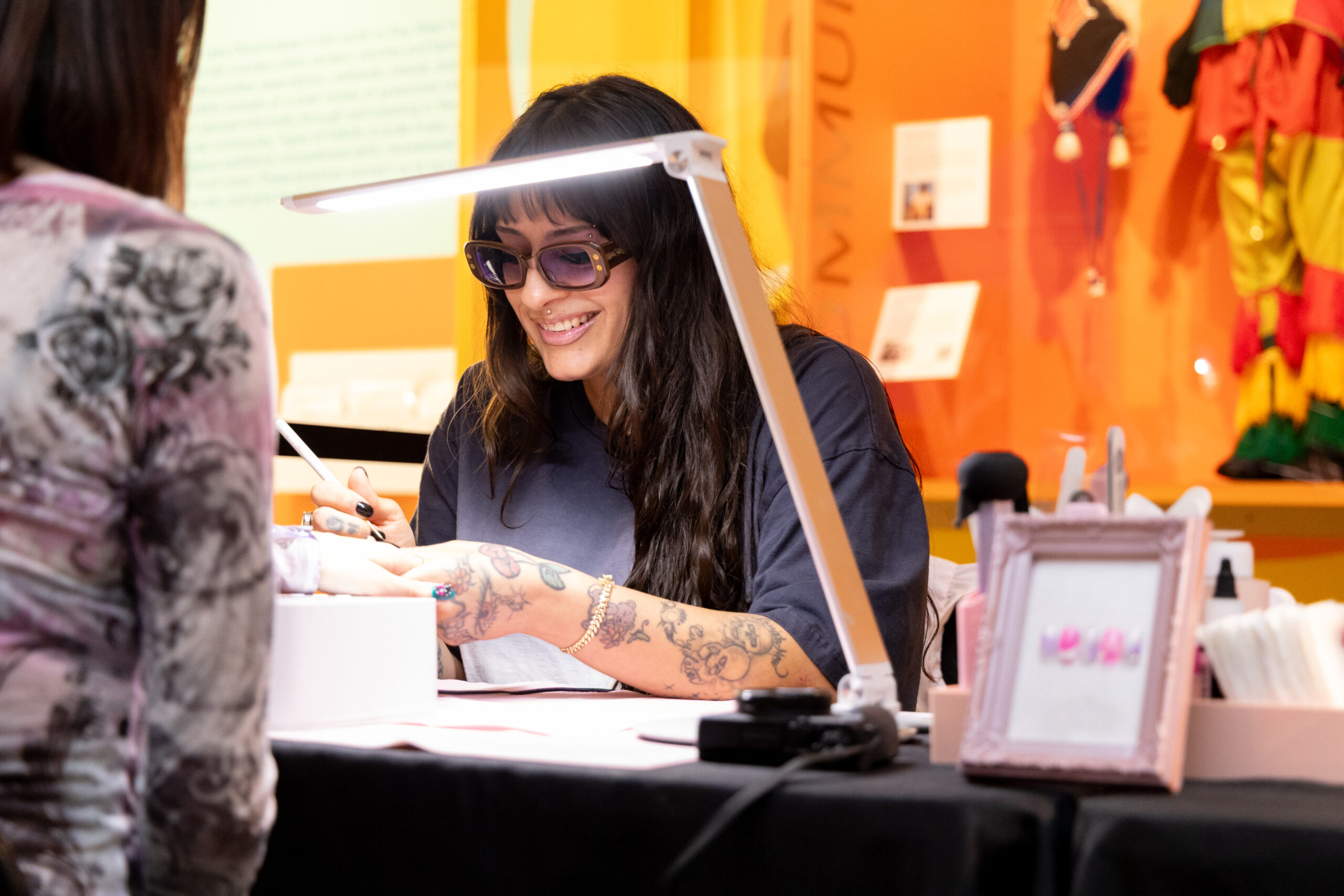 Kro Vargas sits across a table from a client and smiles. There is a work lamp shining over her hands as she holds her client's hand with one hand and a brush in the other.