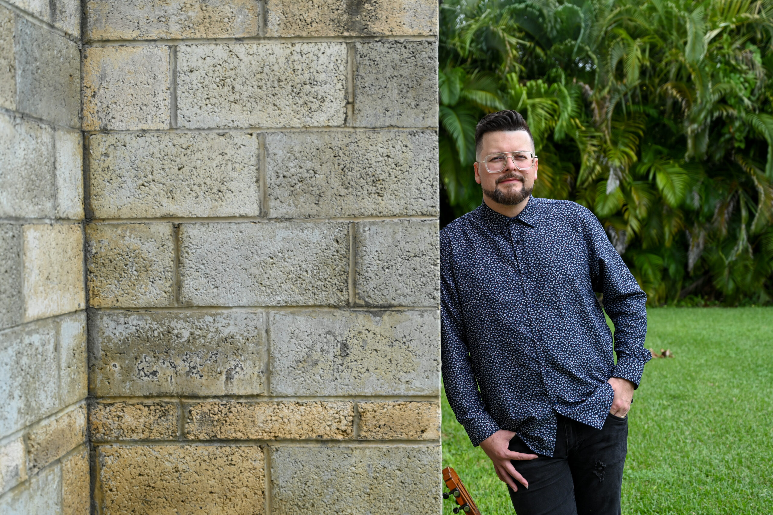 Henry stands leaning on a wall staring at the camera. His cuatro guitar is on grass at his feet, upright and leaning on the wall.