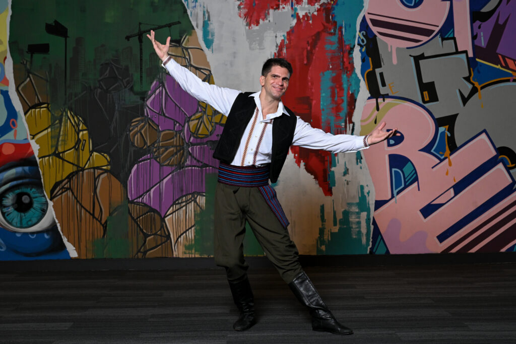 a man stands against a colorful background with his arm spread out and one toe pointed while smiling. He is wearing a white shirt with embroidered designs, a black vest, green pants, and a colorful belt