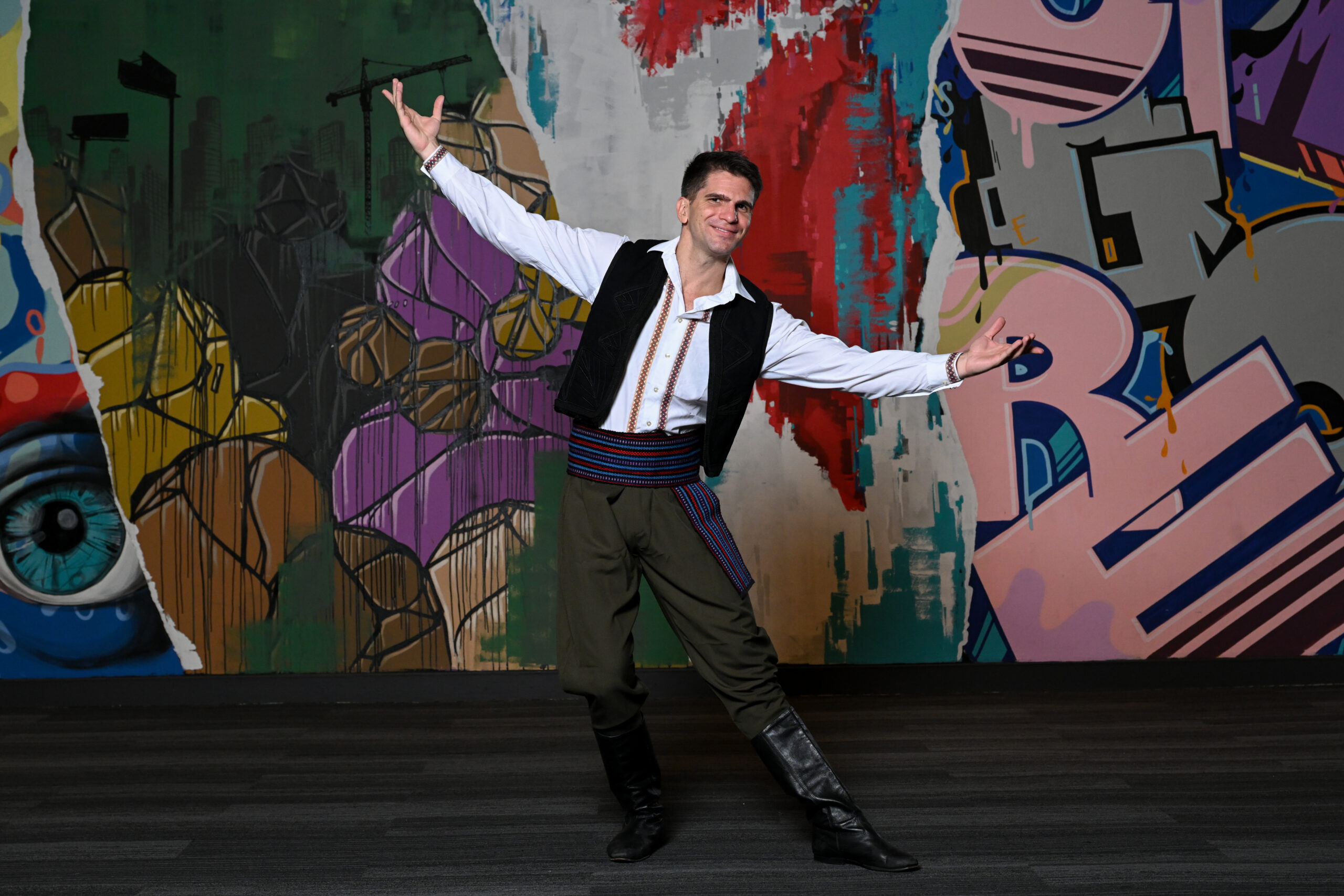 a man stands against a colorful background with his arm spread out and one toe pointed while smiling. He is wearing a white shirt with embroidered designs, a black vest, green pants, and a colorful belt