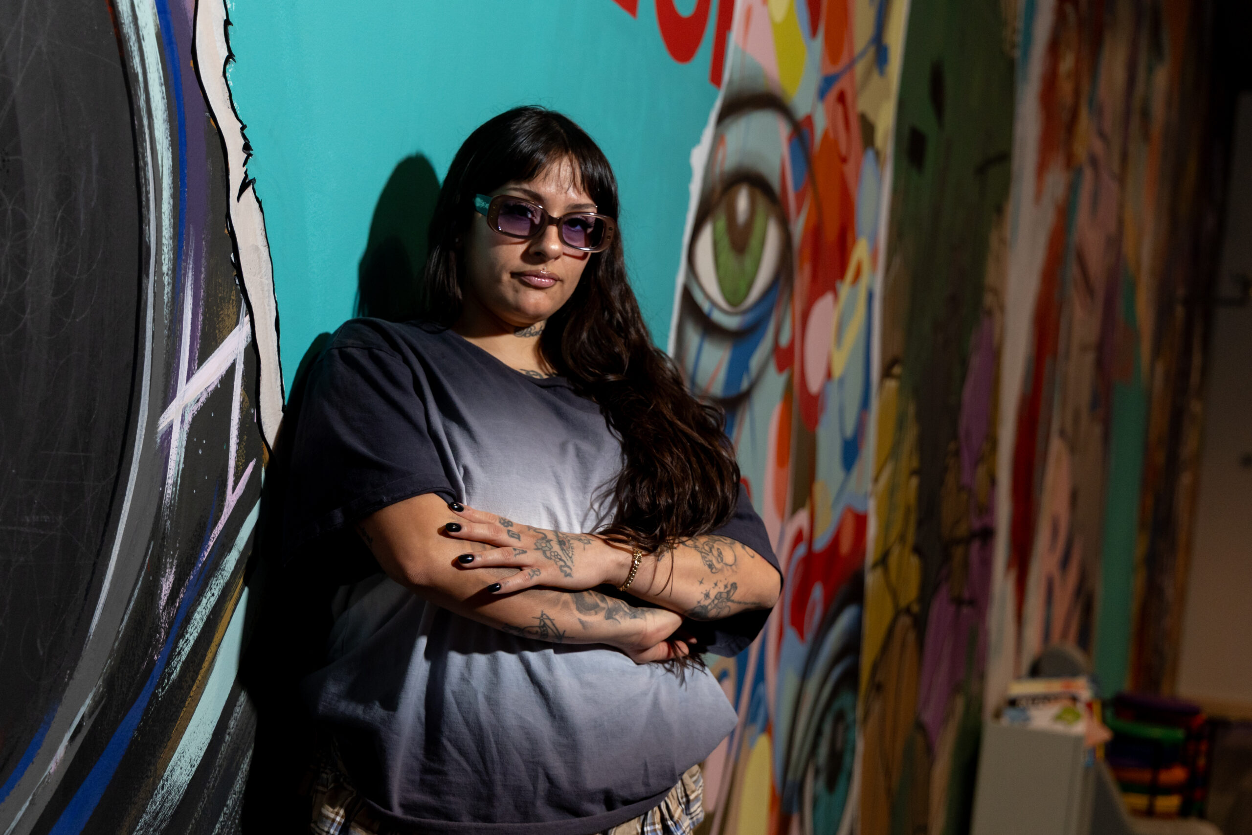 Kro Vargas stands against a wall with her arms crossed, posing for the camera with a serious, confident expression
