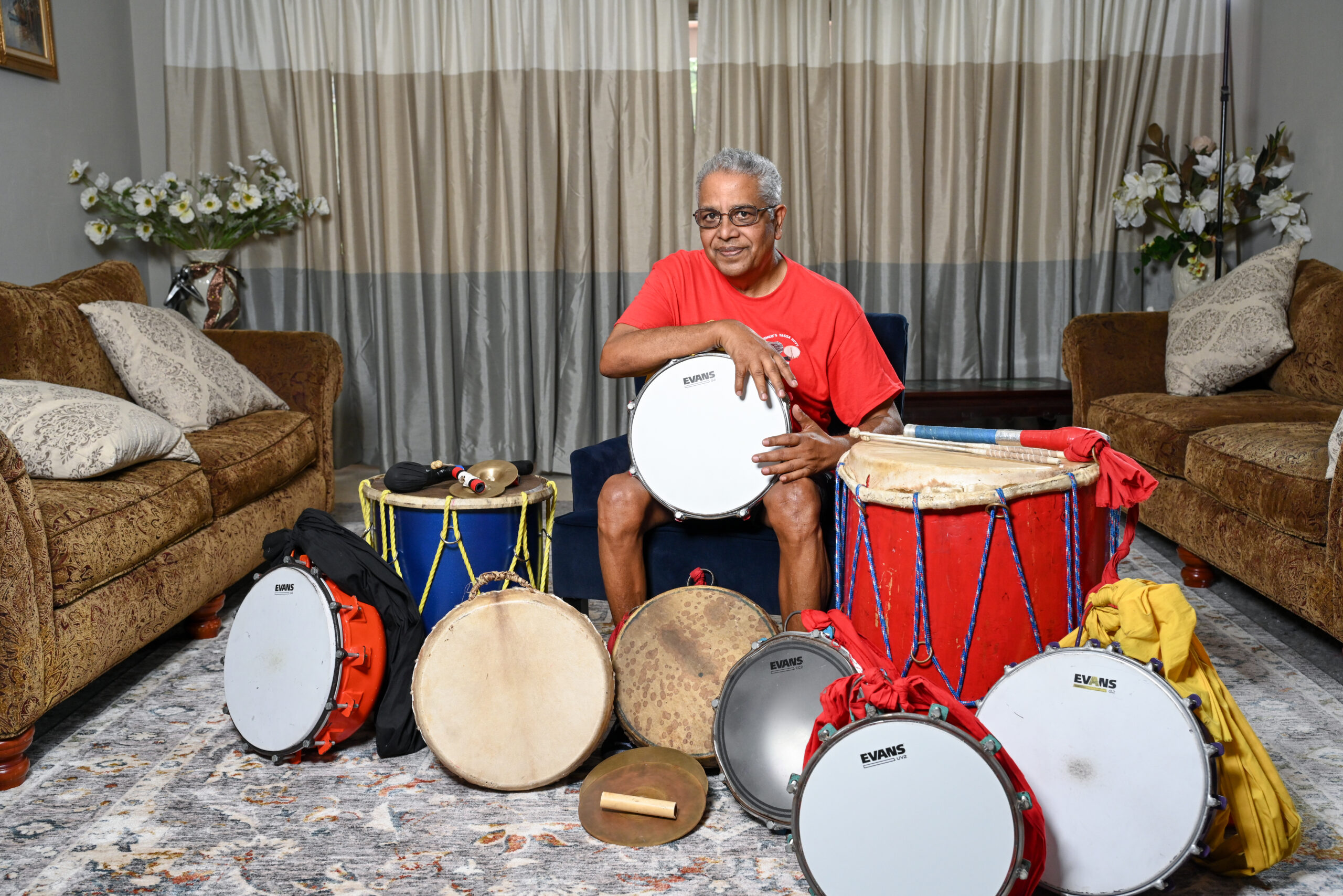 Romeo Ragbir sits surrounded by drums of various sizes. He holds a drum in his hands on his lap.