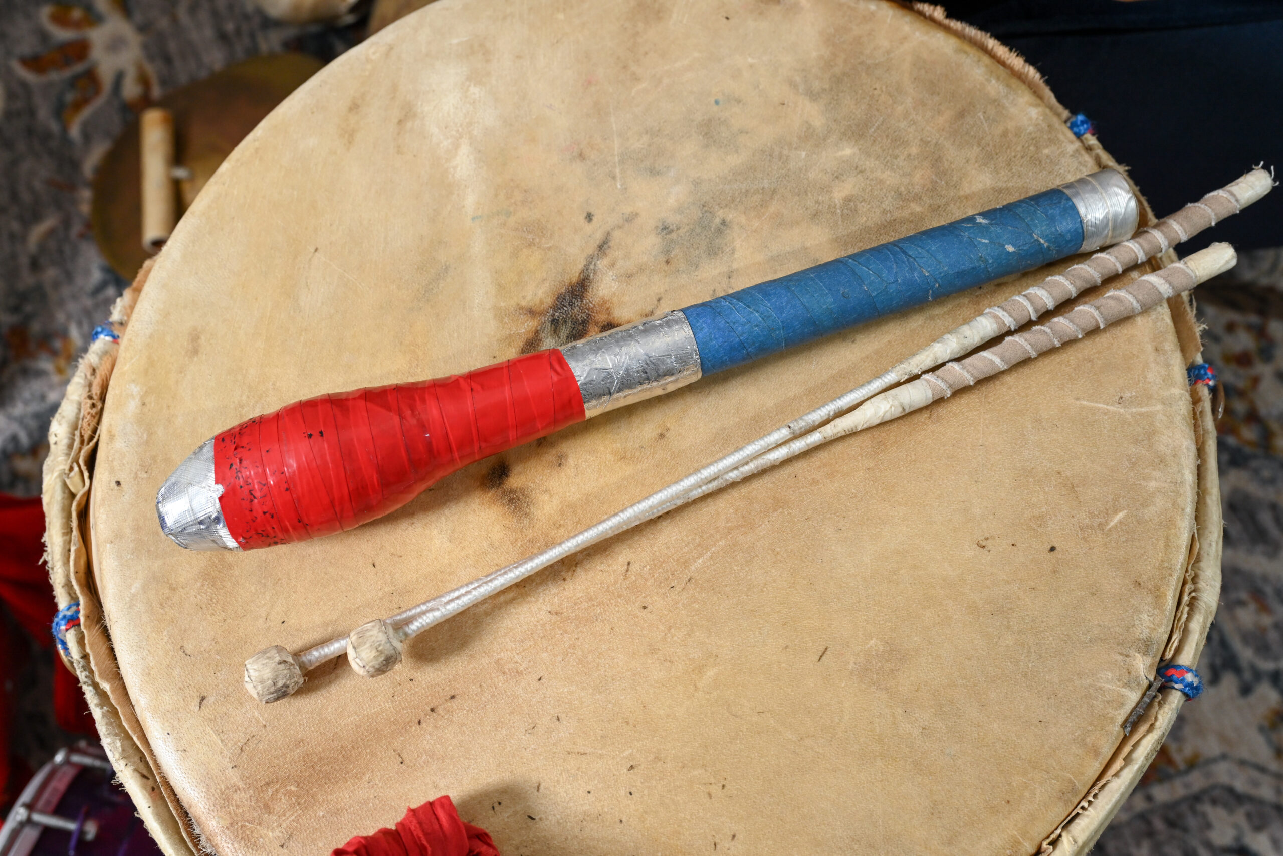 A thick drum stick wrapped in colorful tape sits on a drum head next to a set of thin beige sticks with a round head.