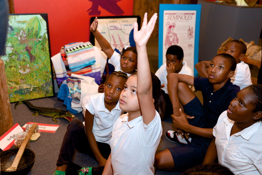 Group of students sitting down, girl in foreground is raising her hand.