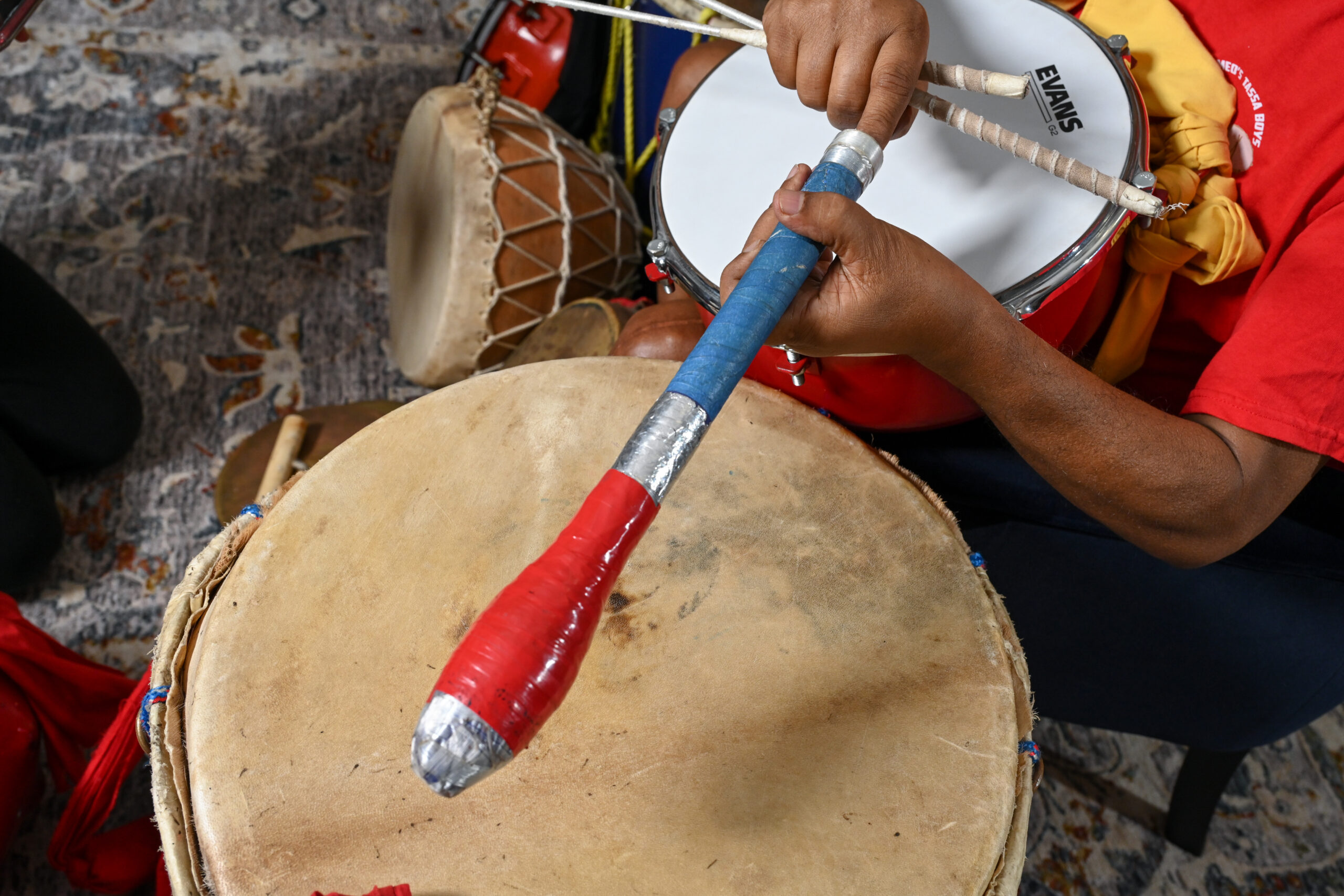 A pair of hands holds a thick drum stick wrapped in colorful tape over a drum head.