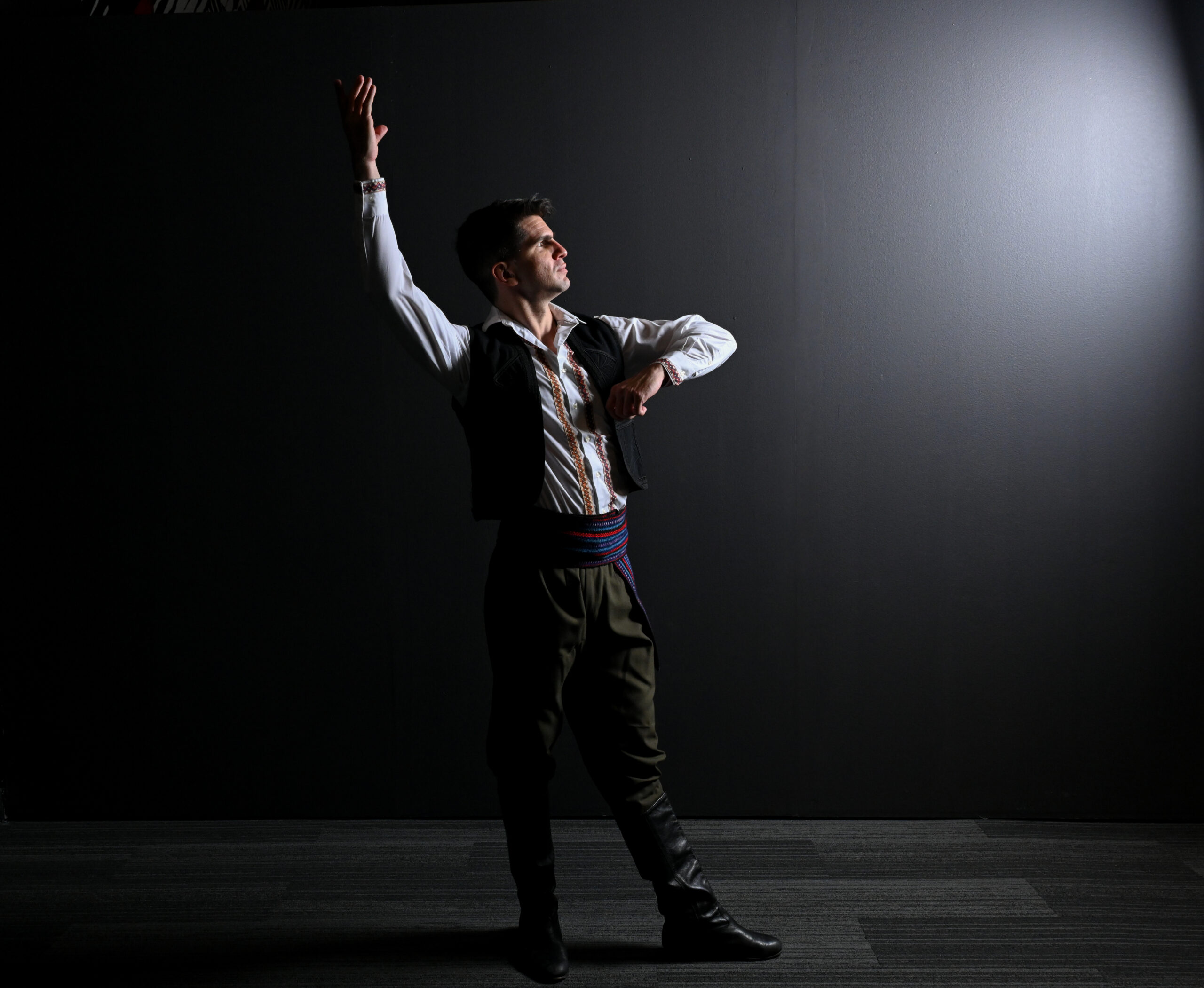 A man stands against a black background with a white light shining on him. He is looking at the light and standing with one arm in the air and one bent at the elbow