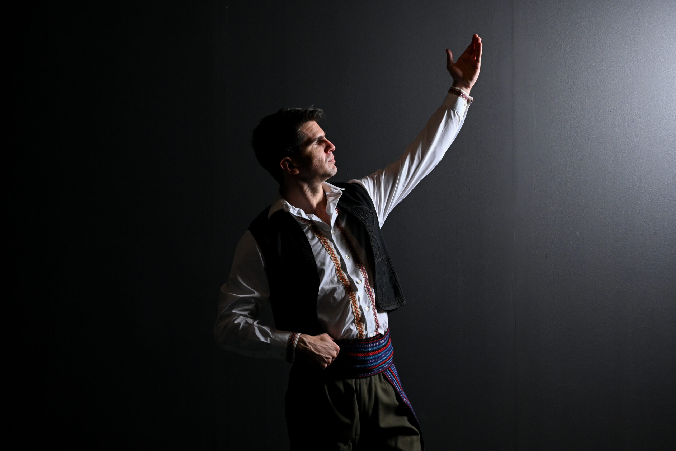 A man stands against a black background with a white light shining on him. He is looking at the light and standing with one arm in the air and one at his waist