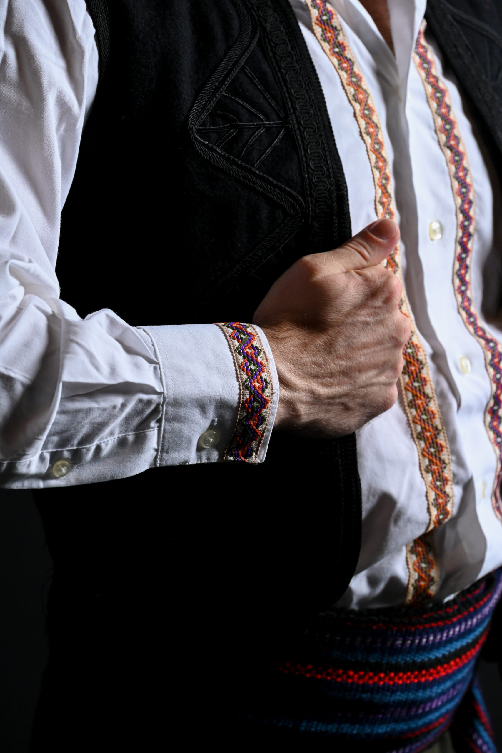 a close up shot of the colorful embroidery along the trim of a white long sleeved shirt and the black stitching on the black vest.