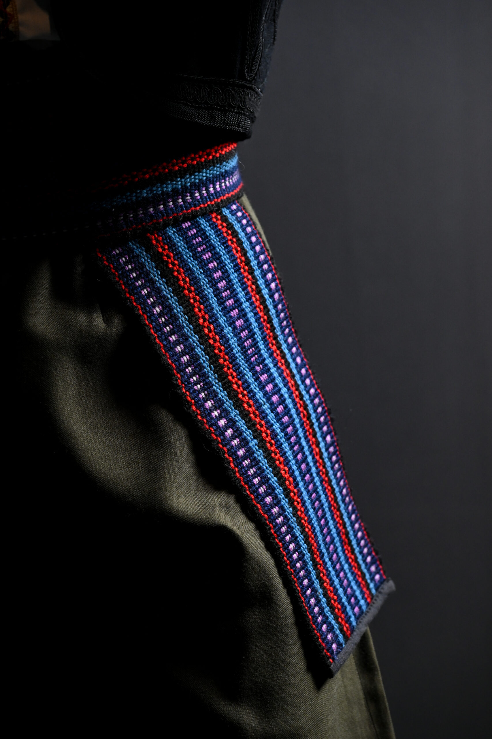 close up shot of the colorful belt with worn around the waist with on end extending down the hip. The belt is made of fabric with patterns in purple, red, blue, and black.