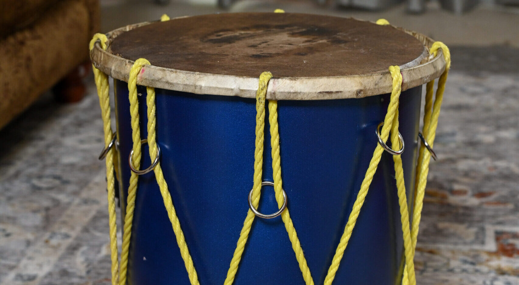 A large, blue drum with a goat skin head sits on the ground. It has yellow strings running up and down its sides to hold the drum head in place.