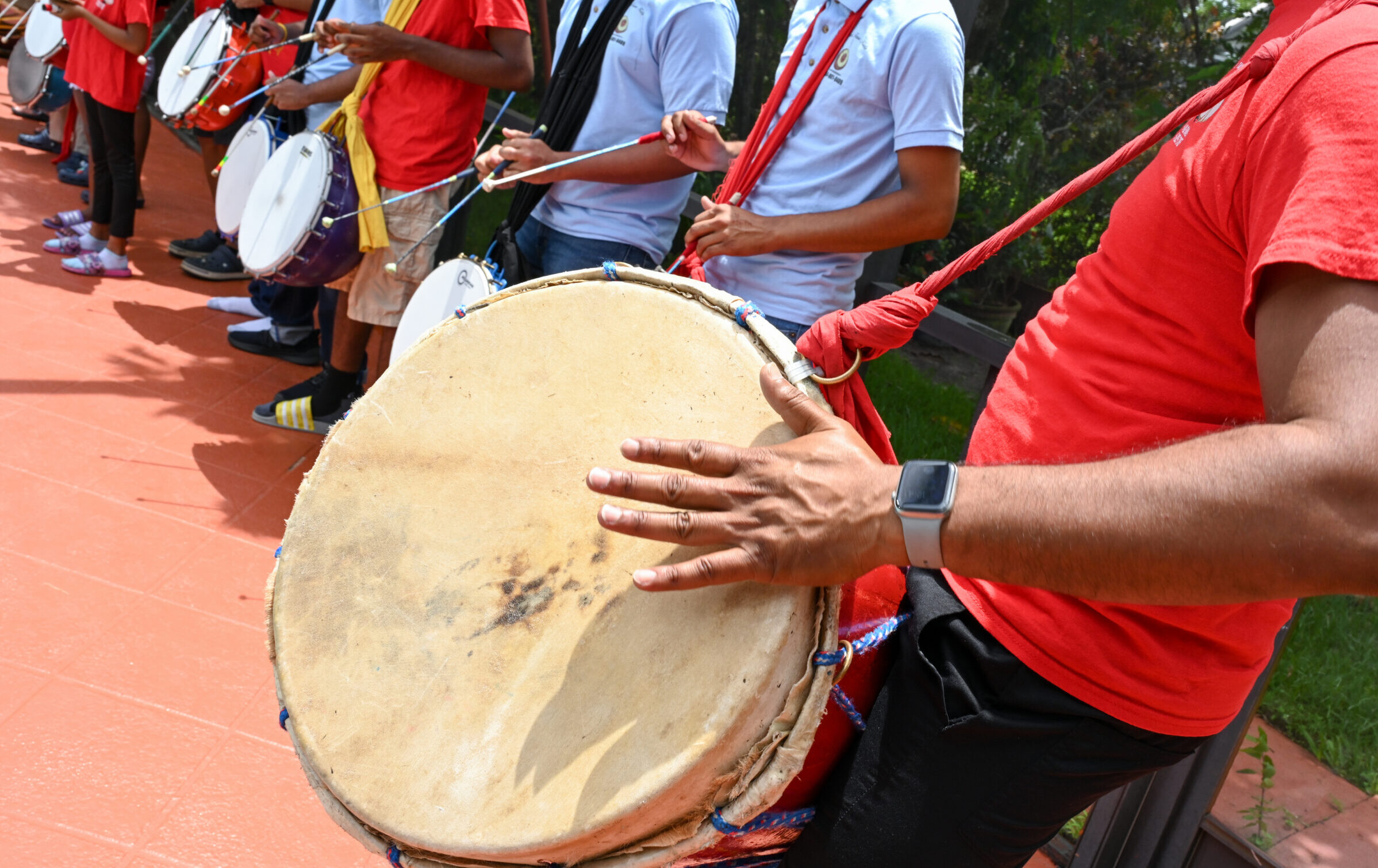 A drummer hits a drum with his hand. In the background, drummers use sticks to hit drums hanging from their necks.