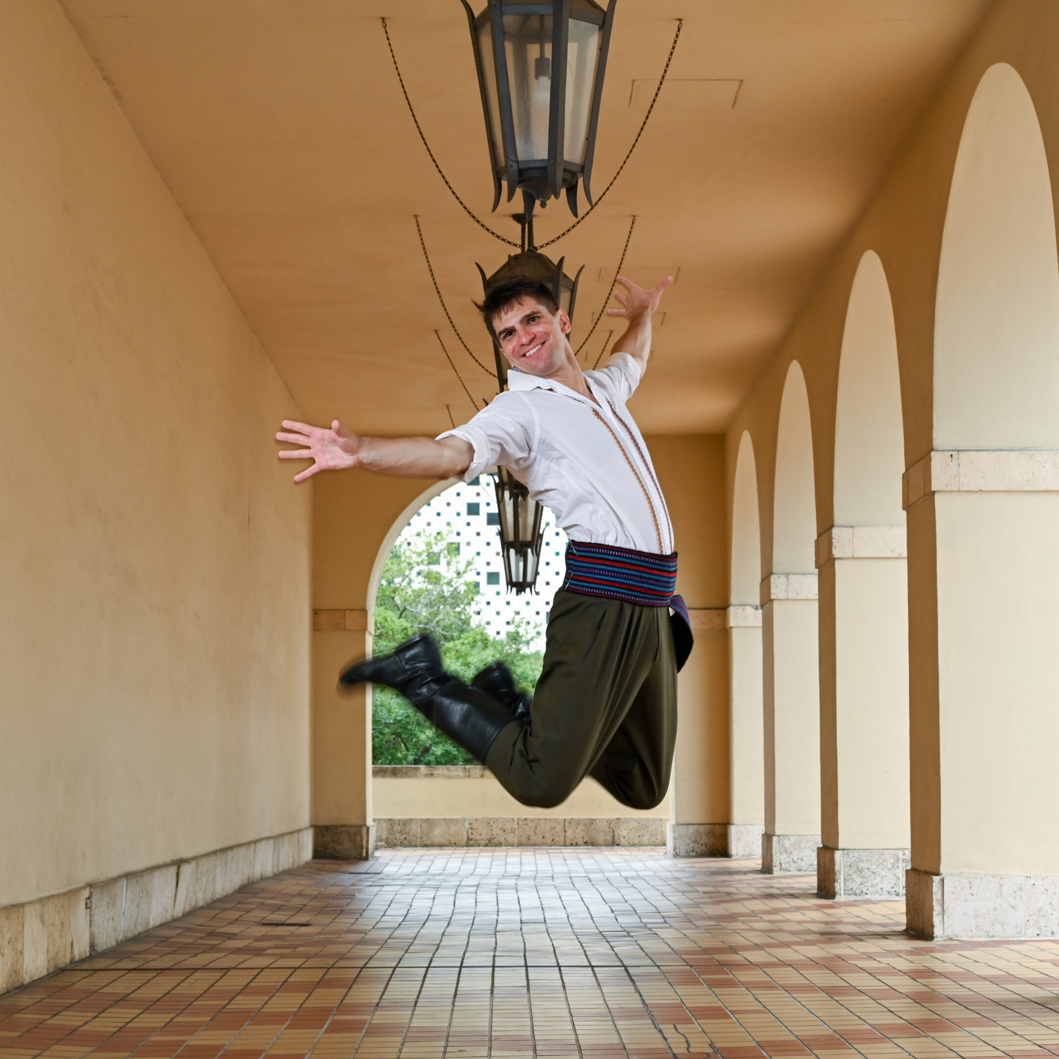 A dancer jumps in the air with his legs and arms stretched behind him. He outdoors in a corridor with hanging lamps and is wearing a white shirt with embroidered designs, green pants, and a colorful belt
