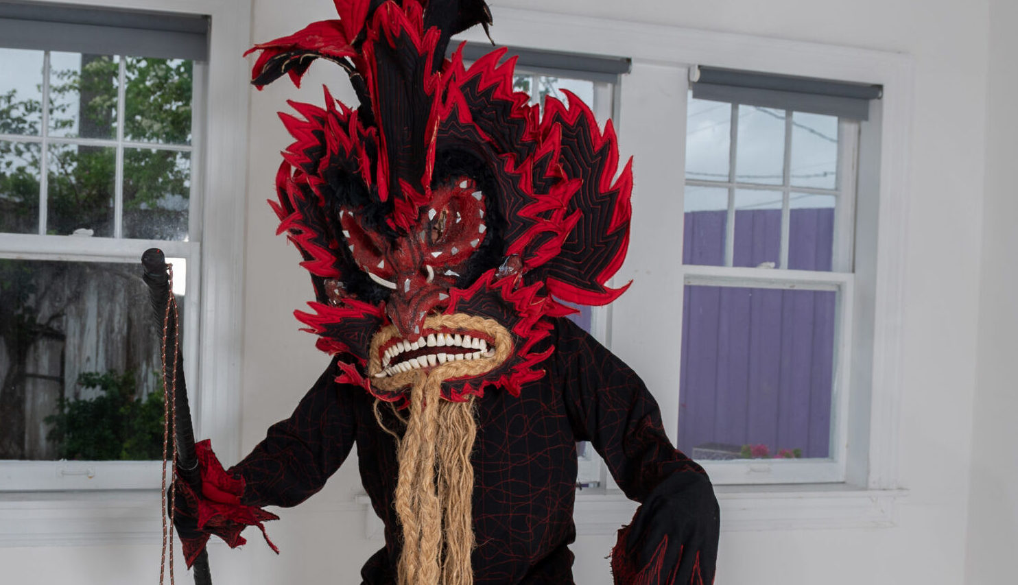 A man in a costume resembling a devil. He wears a red and black mask with large teeth, red eyes, and black and red fabric fanned around the face. Straw material hangs from the bottom of the mask resembling a beard. Black and red feathers stick out of the top. His body suit is black with red accents and he holds a black stick up in one hand.