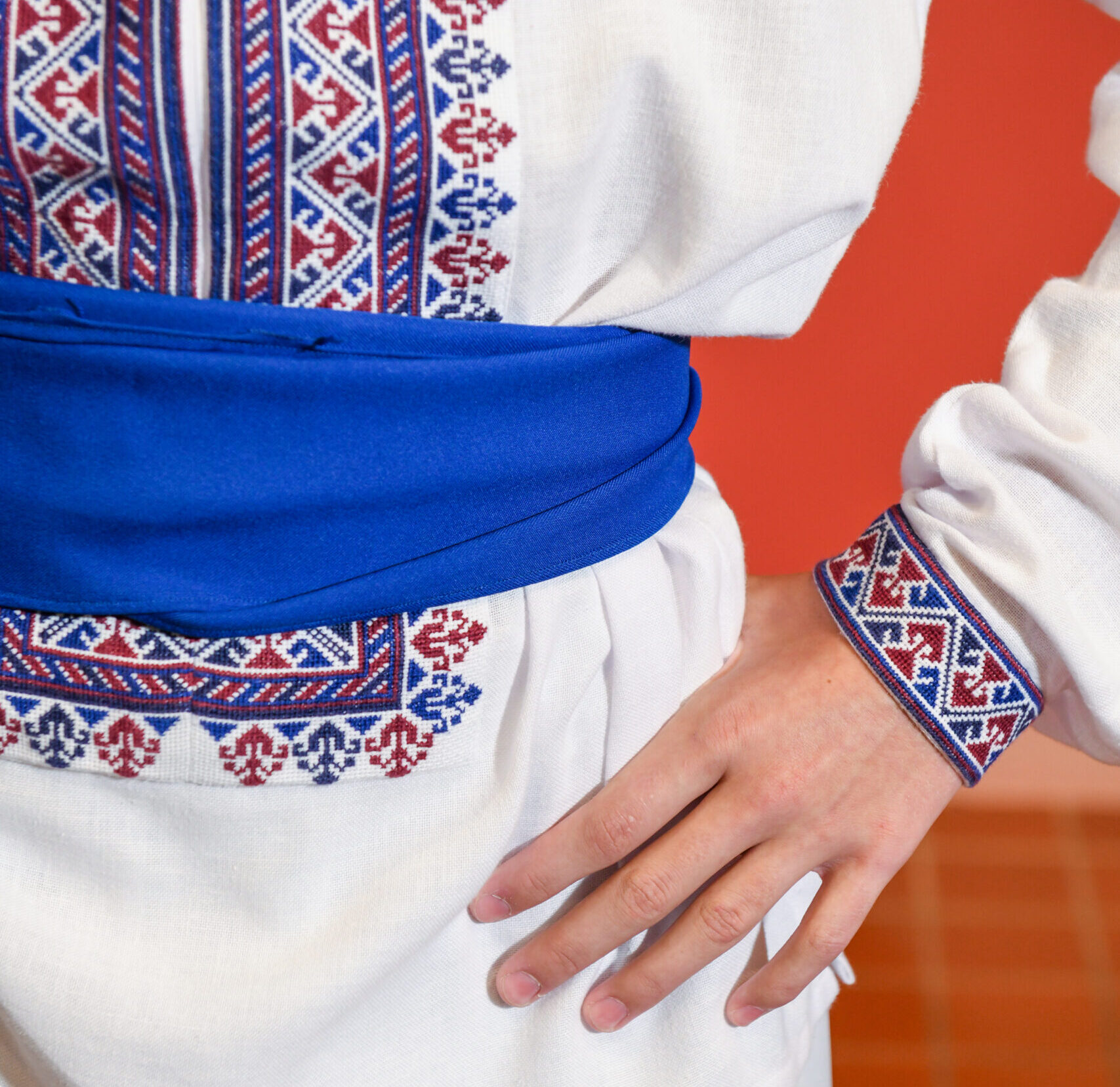 Details of a male dancers costume including a blue sash around the waist and blue and red embroidery on the front of the shirt and the sleeve.