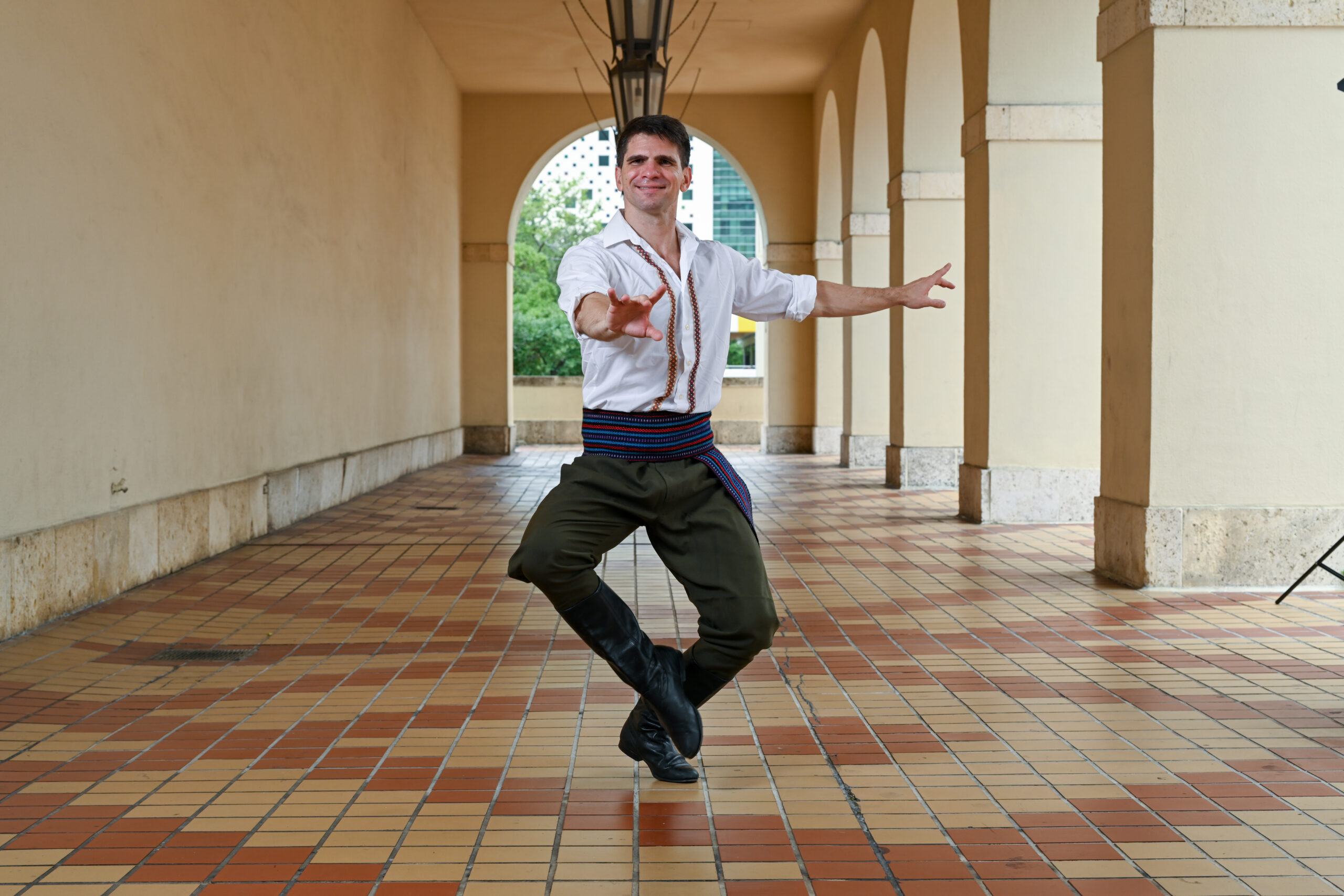 A dancer poses with his toes pointed towards the center and his arms outstretched. He outdoors in a corridor with hanging lamps and is wearing a white shirt with embroidered designs, green pants, and a colorful belt
