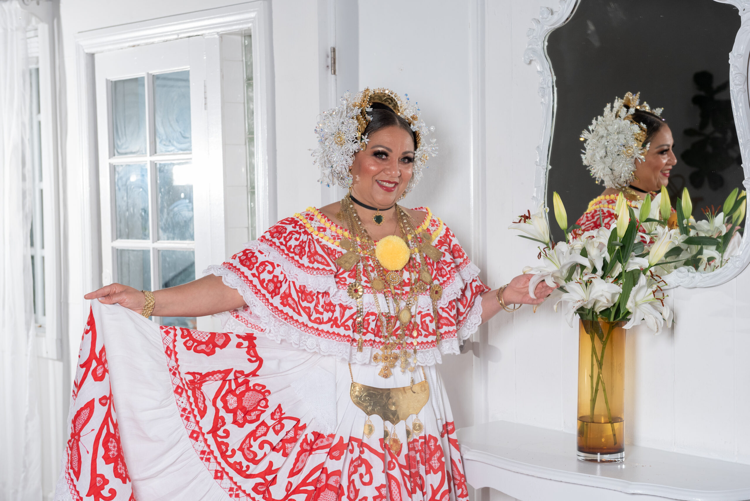 A woman wears am elaborate embroidered red and white dress with yellow accents. She wears a white beaded headpiece and gold chains. She is posing next to a vase of flowers.