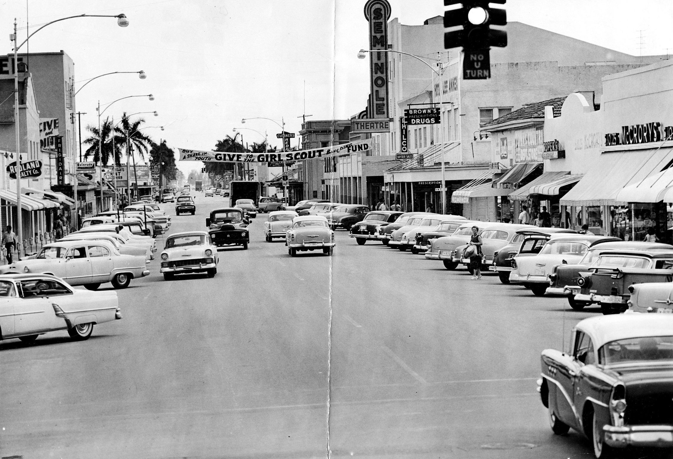 View looking south on Krome Avenue. Krome Avenue is featured in the middle of the photograph with a view of cars parked on either side of the street. The Seminole Theatre sign can be seen atop a building in the background.