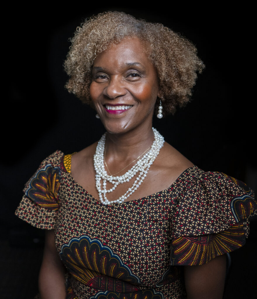 A person with short curly brown and grey hair sits before a black background. They're smiling and wearing white dangly pearl earrings, a white multi-layered beaded necklace, and a colorful patterned blouse with ruffled shirt sleeves.