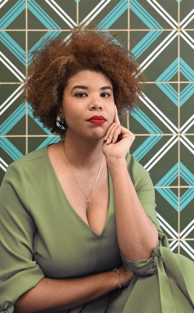 A person with brown skin and light brown curly short hair sits with an arm across her middle and her chin resting on her other hand. They look directly towards the camera, wearing a green top, against a patterned wall.