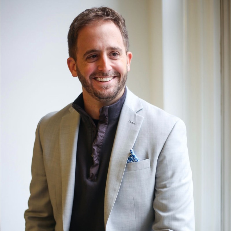 A person with a light brown mustache and beard smiles and glances away from the camera. They wear a great suit and dark colored shirt.