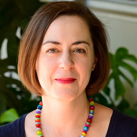 A person with short chin length brown hair smiles at the camera. They wear a brightly colored beaded necklace and navy top.