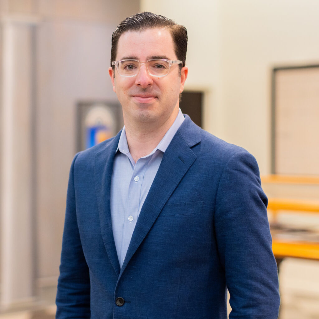 A person with short brown hair and clear rimmed glasses smiles at the camera. They wear a blue suit and pale blue button down shirt.