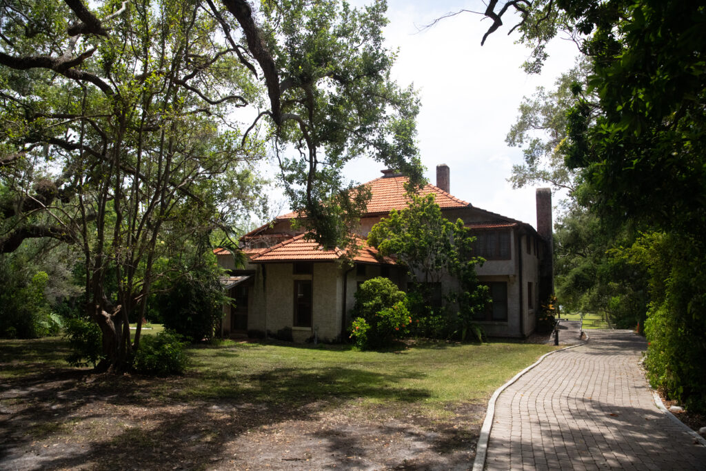View of The Barnacle house, a two-story beige house with orange roof tile. Trees can be seen in the foreground. 