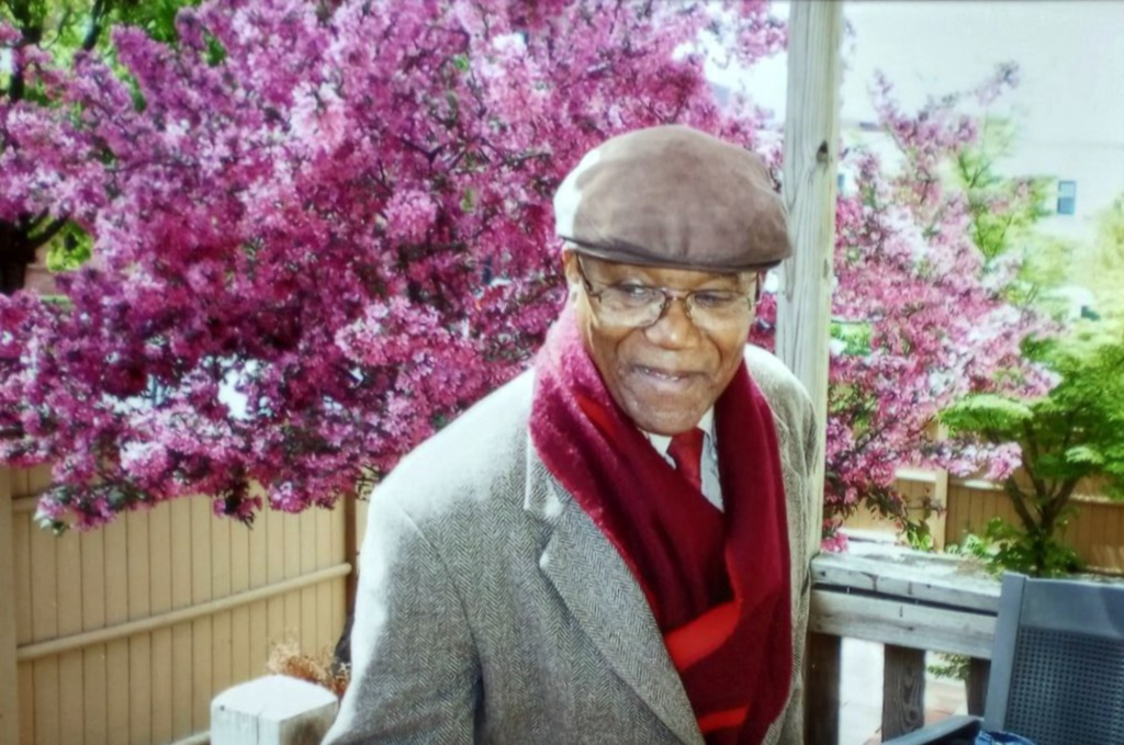 A person wearing a brown cap, light brown wool suit jacket, and a red scarf and tie stands looking away from the camera. 