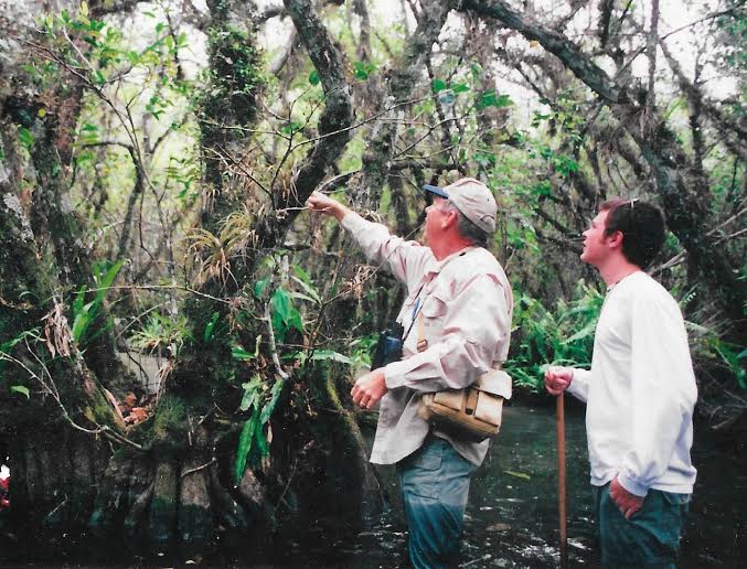 Two men, one younger and one older, are looking at a tree in a lush, overgrown area. The older man points at the tree in the direction both men are looking.