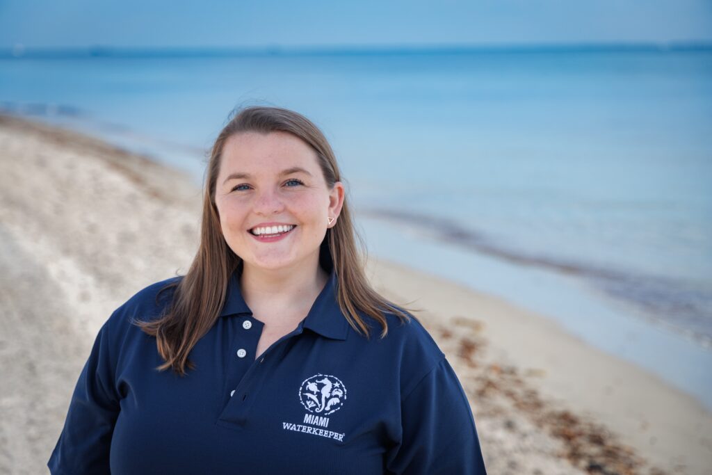 A person with mid-length mousy brown hair smiles for a photo along a beach with light blue waters. They wear a navy blue polo with a logo and the words, "Miami Waterkeeper."