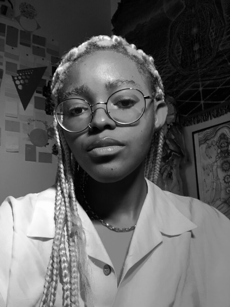 Black and white headshot of a person wearing glasses and a lightly colored collared shirt. 2D art is seen hanging on the wall in the background.