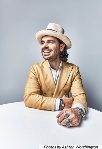 Gabriel Urrutia wears a yellow suit jacket, white shirt, and beige hat. Gabriel smiles as he looks in the distance.