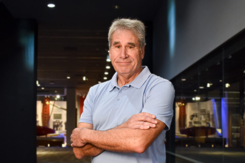 A person wearing a collared blue shirt stands with their arms crossed in front of a museum exhibition.