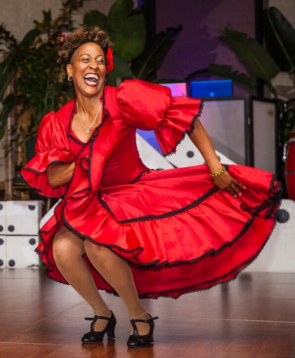 Dancer in a red and black dress smiling as she moves her skirt with her arms.