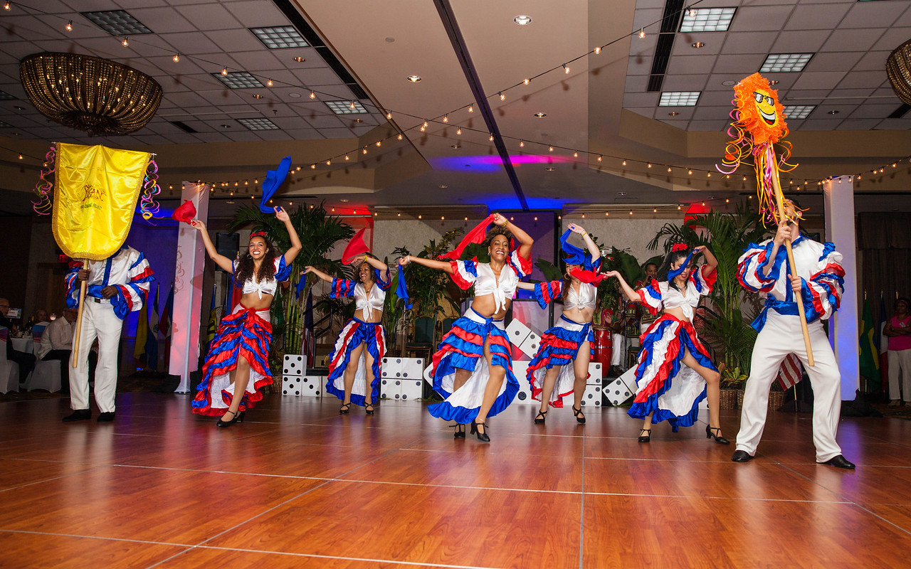 Five women dancers wear red, white, and blue skirts and tops and waive red and blue scarves in the air as they dance.