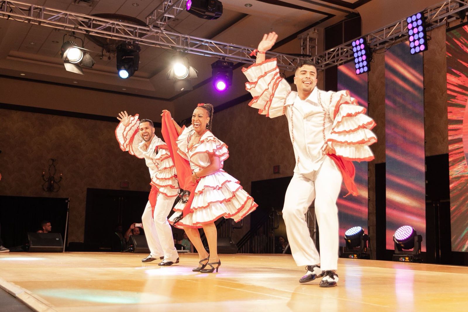 two male dancers and a female dancer dance in red and white costumes. The men wear puffy sleeves and the woman carries a red scarf in her hands.