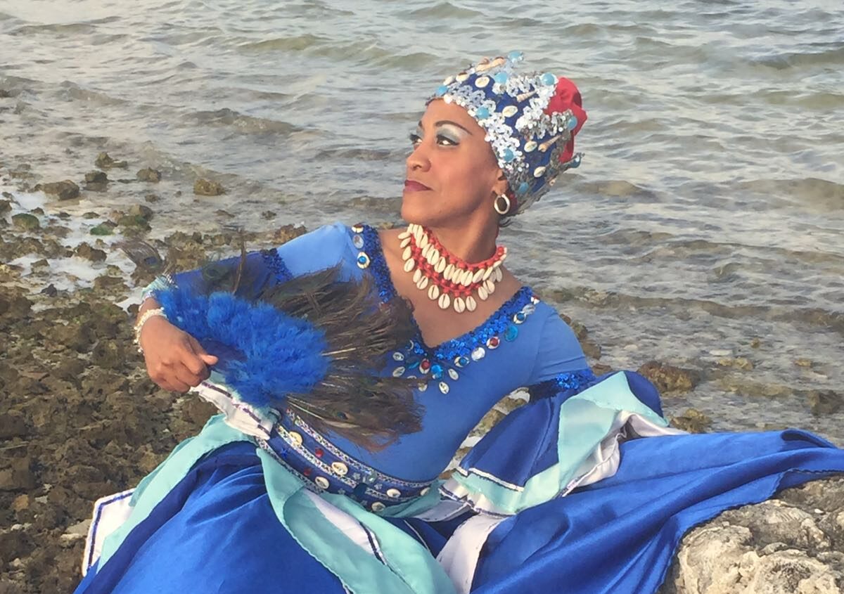 A woman in a blue dress holds a fan with peacock feathers. She is sitting by the ocean.