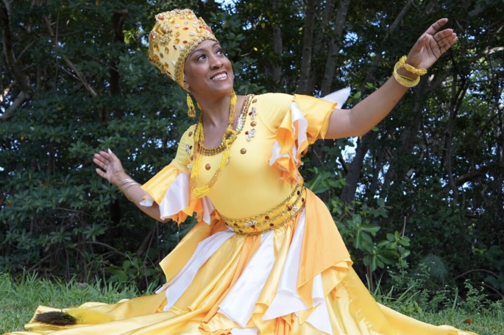 A woman poses with her arms lifted wearing a yellow crown and dress. She is posing in front of trees.
