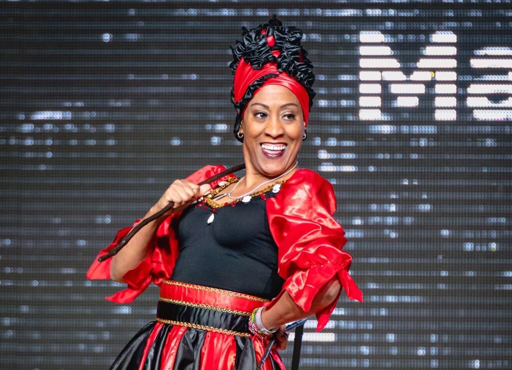 a woman poses in a red and black outfit. She wears a turban style headscarf and carries a thin stick over her shoulder