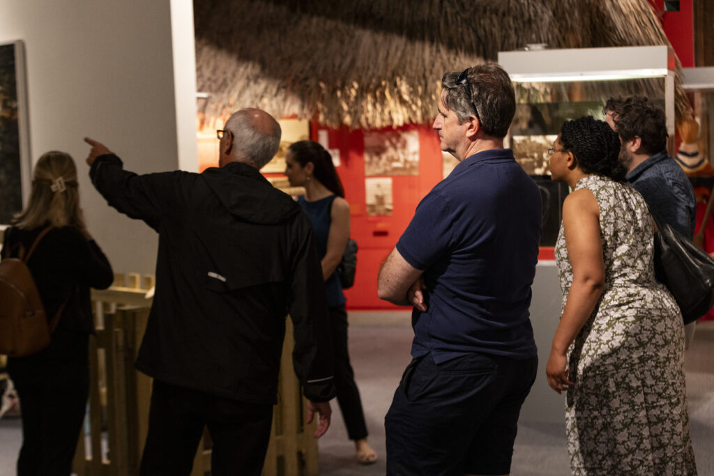 A person points to an object inside of a Museum exhibition to a group of adults who look on.