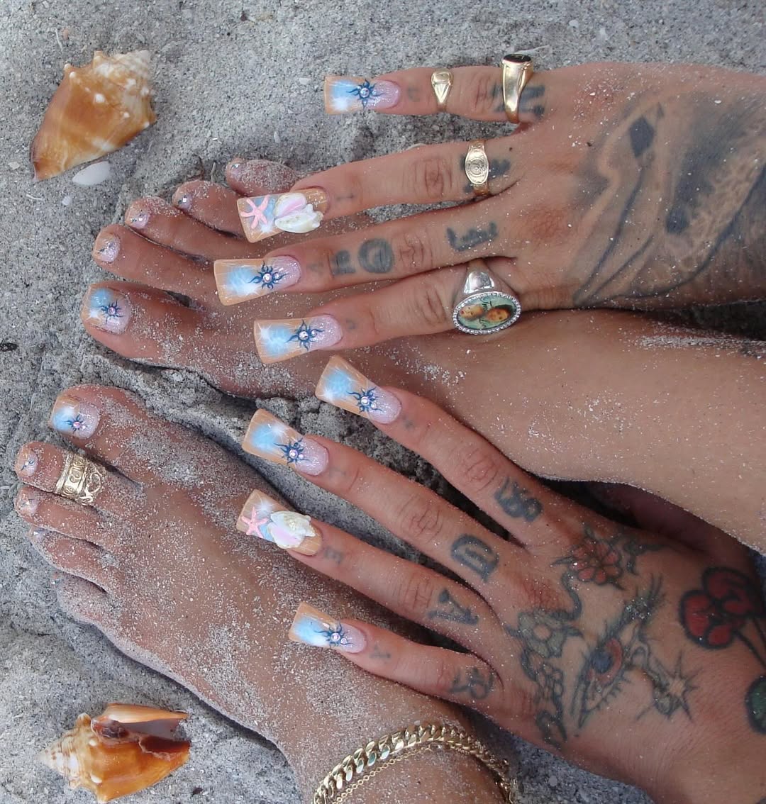 hands and feet are in the sand with a blue, coral, and pink nail design that features conch shell sculptures and blue sun designs with rhinestones in the middle