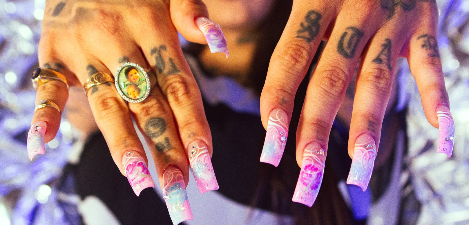 A close up of hands with the focus on the purple. pink, and blue design of her nails, her jewelery, and the tattoos on her fingers and the back of her hands