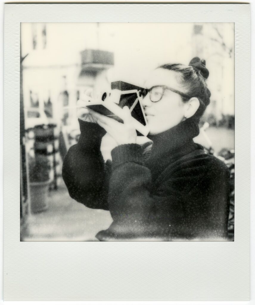 A black and white image of a person wearing their hair in a top knot and thick rimmed glasses with a turtleneck sweater. They hold up a vintage camera to one eye as they look through it.