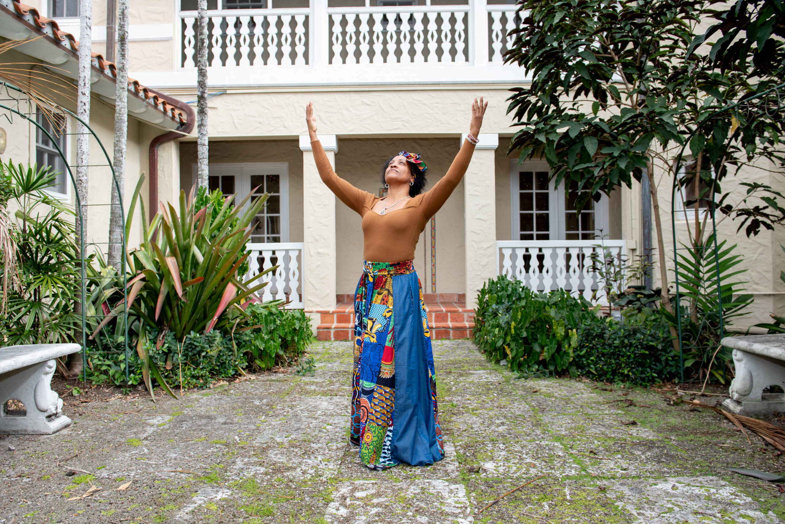 A woman stands in a garden with her arms lifted and looking up at the sky