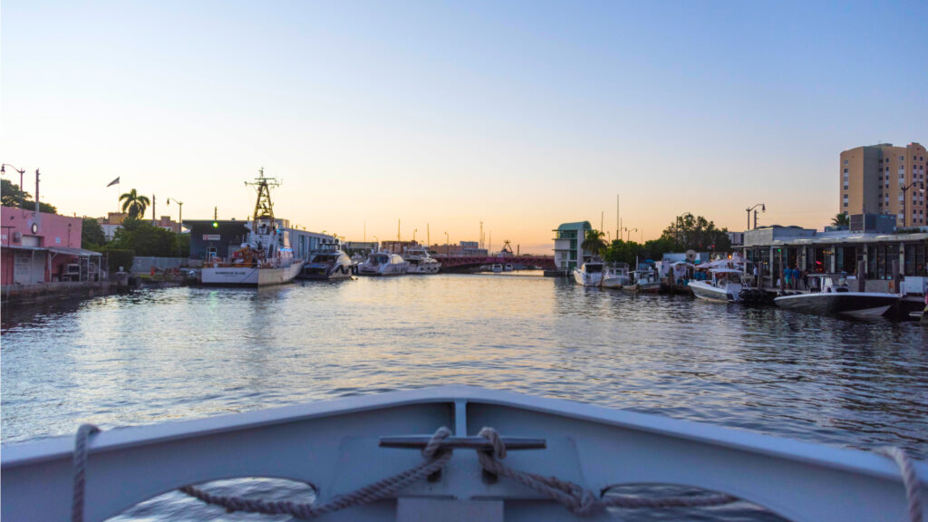 View of the Miami River at dusk from the front of a boat. The sunset is reflected upon relatively still water with boats docked along the shoreline.