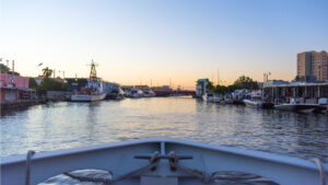 View of the Miami River at dusk from the front of a boat. The sunset is reflected upon relatively still water with boats docked along the shoreline.