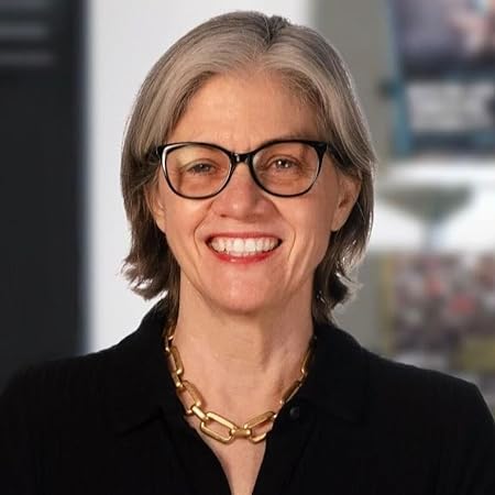 A person with silver shoulder length hair wearing black rimmed glasses smiles at the camera. They wear a black collared shirt and gold chain necklace.