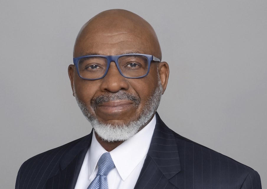 A person wearing thick rimmed glasses and a suit and tie stands in front of a grey background.