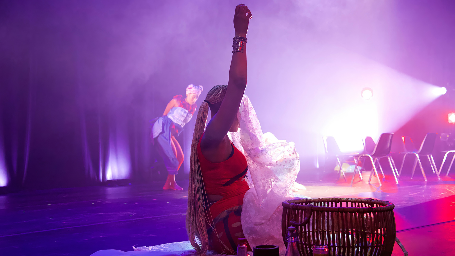 A person sits on a stage with one arm raised. They wear an orange shirt and have long blonde hair tied in a ponytail. A white cloth covering shrouds their face as they look away from the camera. They are surrounded by two woven baskets, glass bottles, and tall pillar candles in glass containers. A person can be seen in the distance behind them mid-movement on stage.