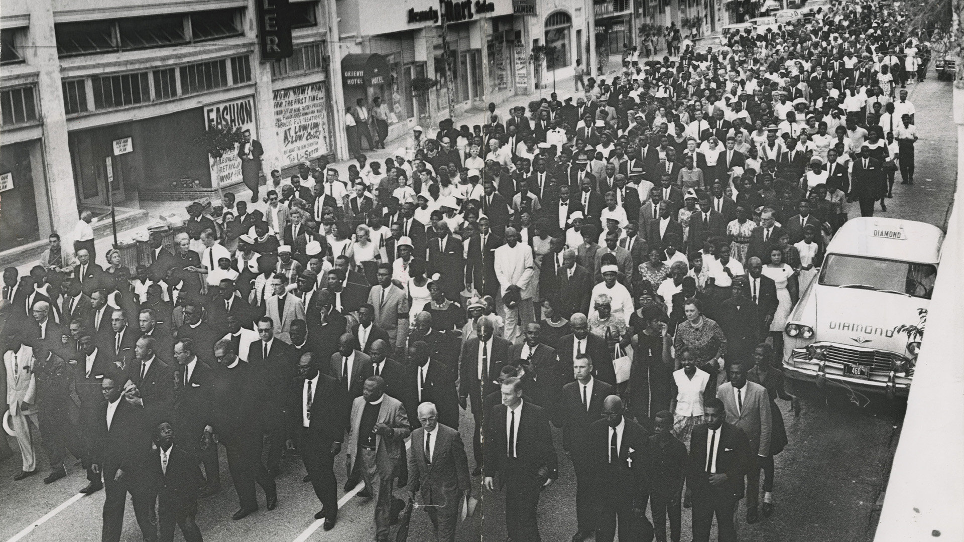 A black and white archival image. A crowd of people march through the streets of Miami in protest.